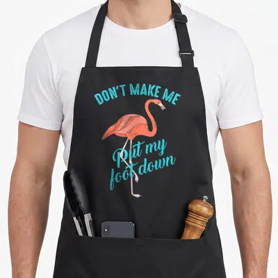 Foot flamingo apron for him - Pink cartoon flamingo with cyan text reading DON'T MAKE ME Put my foot down — shown on a black cotton canvas apron against a white background