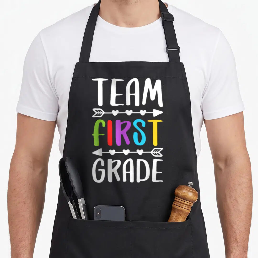 Grade 1st teacher apron - Stacked text reading TEAM FIRST GRADE with colorful letters and heart arrows — shown on a black cotton canvas apron against a white background