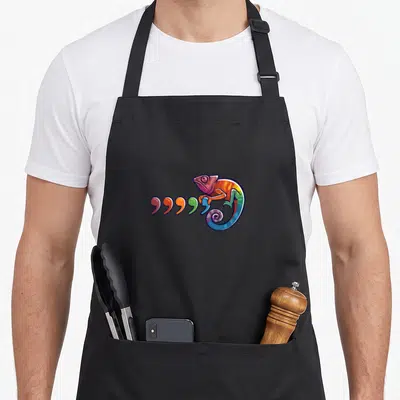 Chameleon 80s apron - Cartoon chameleon curled in spiral next to five colored commas in pink, orange, yellow, green — shown on a black cotton canvas apron against a white background