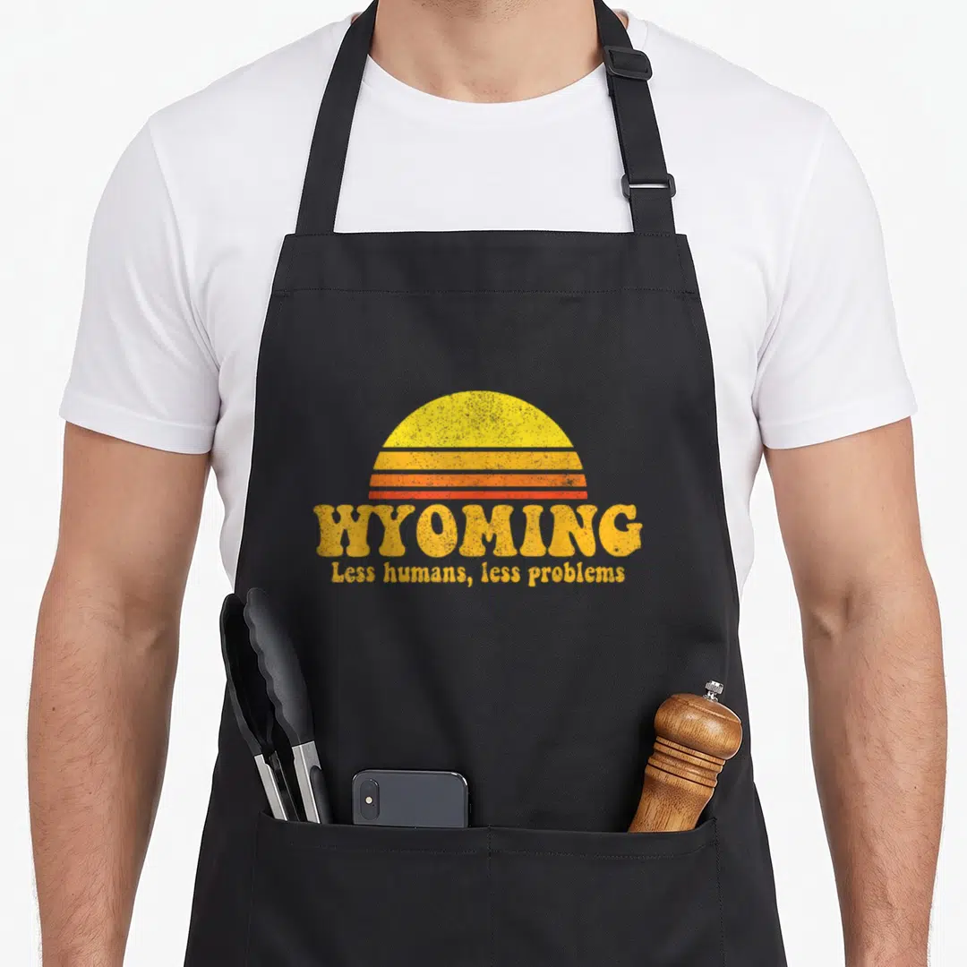 Wyoming us aprons - Retro sun with yellow orange red stripes and text WYOMING Less humans less problems — shown on a black cotton canvas apron against a white background