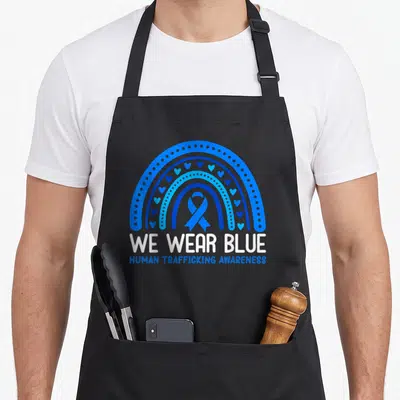 January rainbow apron - Blue rainbow with hearts and dots arching over We Wear Blue text and awareness ribbon — shown on a black cotton canvas apron against a white background