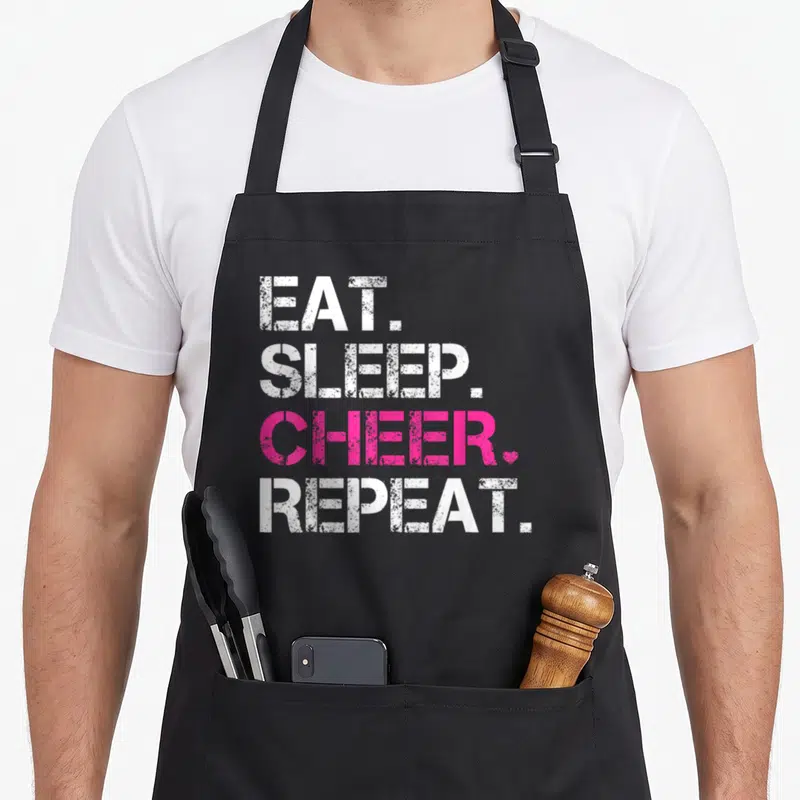 Eat cheers years apron - Stacked EAT SLEEP CHEER REPEAT text in white and pink with a small heart icon — shown on a black cotton canvas apron against a white background