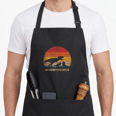 Dinosaur mommy daughter apron - MOMMYSAURUS text under large and two small dinosaur silhouettes on striped sun — shown on a black cotton canvas apron against a white background