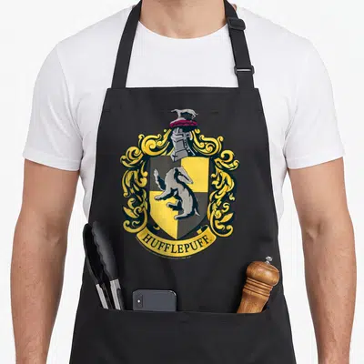 Crest hufflepuff apron - HUFFLEPUFF text below a shield containing a badger and yellow scrollwork — shown on a black cotton canvas apron against a white background