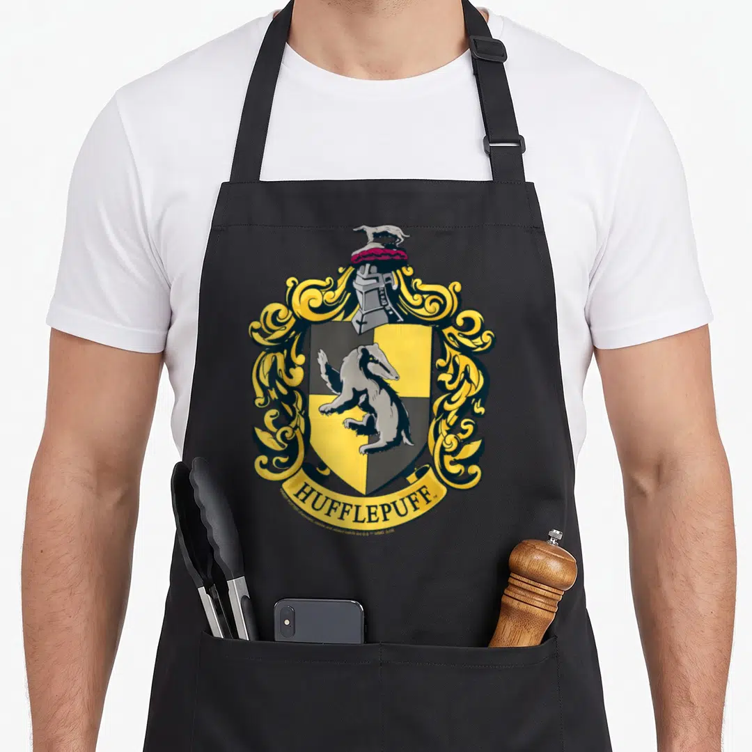 Crest hufflepuff apron - HUFFLEPUFF text below a shield containing a badger and yellow scrollwork — shown on a black cotton canvas apron against a white background