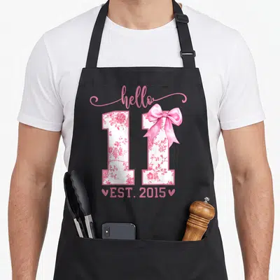 Coquette birthday apron - Pink floral number 11 with bow and EST 2015 text — shown on a black cotton canvas apron against a white background