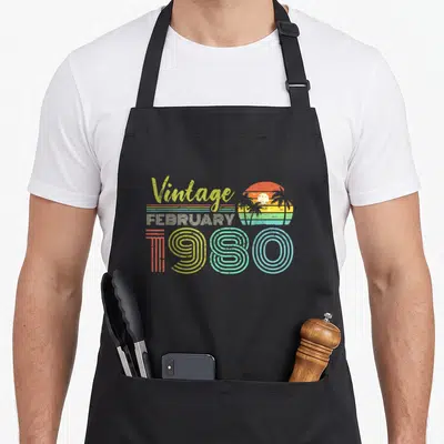 February 40th birthday apron - Vintage FEBRUARY 1980 retro sunset palm tree graphic in orange yellow green blue — shown on a black cotton canvas apron against a white background