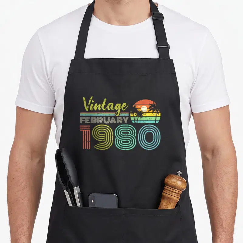 February 40th birthday apron - Vintage FEBRUARY 1980 retro sunset palm tree graphic in orange yellow green blue — shown on a black cotton canvas apron against a white background