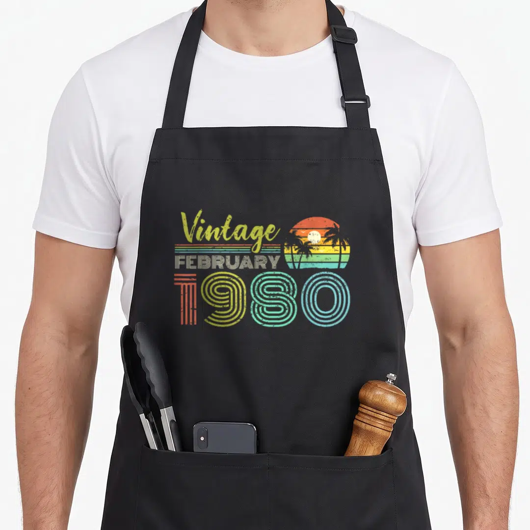 February 40th birthday apron - Vintage FEBRUARY 1980 retro sunset palm tree graphic in orange yellow green blue — shown on a black cotton canvas apron against a white background