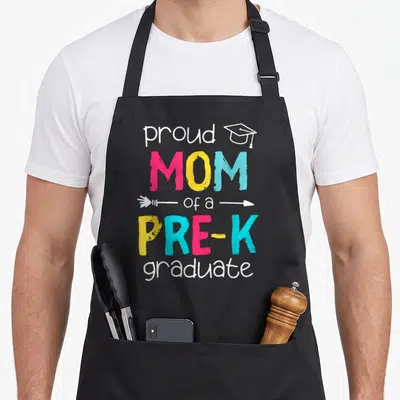 Prek mother's apron - Stacked colorful text reading proud MOM of a PRE-K graduate with cap and arrow graphics — shown on a black cotton canvas apron against a white background