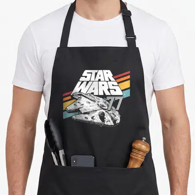 Stripe falcon apron - Star Wars 77 text over Millennium Falcon with red orange yellow teal stripes — shown on a black cotton canvas apron against a white background