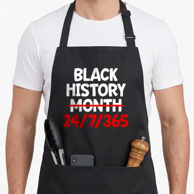 History african print apron - White text reading Black History Month struck through with red line above red 24/7/365 — shown on a black cotton canvas apron against a white background