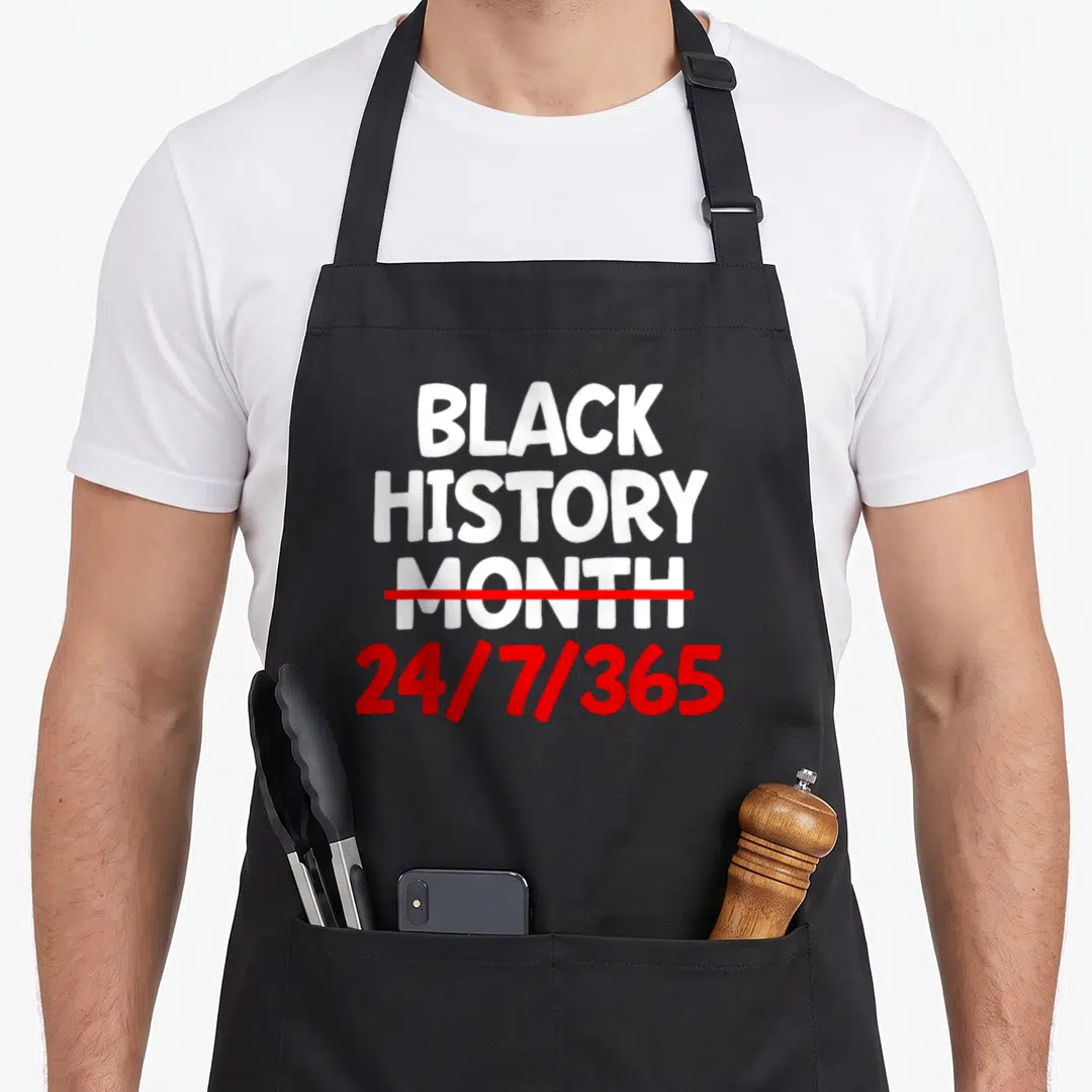 History african print apron - White text reading Black History Month struck through with red line above red 24/7/365 — shown on a black cotton canvas apron against a white background