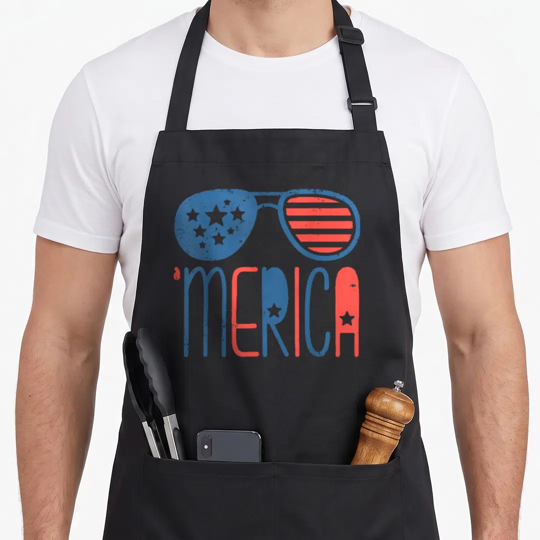 Aviator 4th of july apron - Sunglasses with stars and stripes above 'MERICA text in blue and red — shown on a black cotton canvas apron against a white background