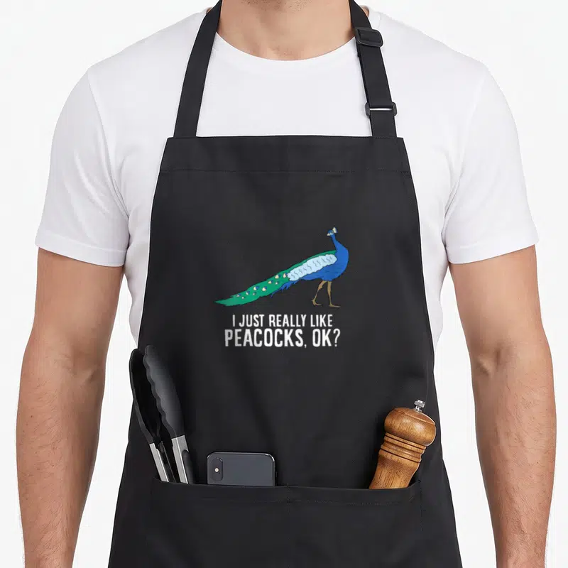 Retro bird apron - Cartoon peacock in blue and green above white text reading I JUST REALLY LIKE PEACOCKS OK — shown on a black cotton canvas apron against a white background