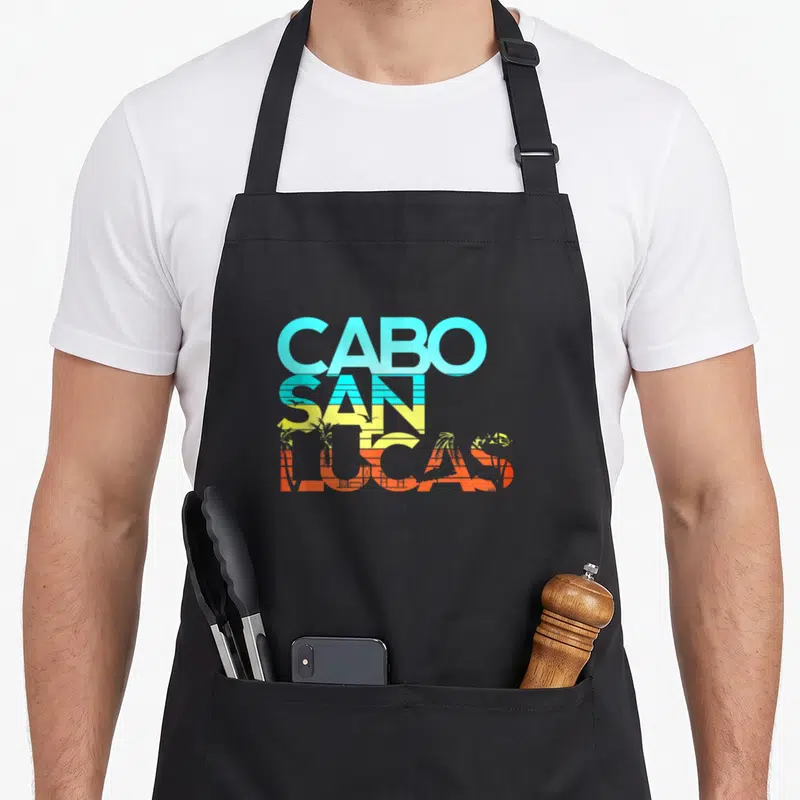 Resort mexico apron - Stacked CABO SAN LUCAS text in blue yellow orange with palm tree silhouettes — shown on a black cotton canvas apron against a white background