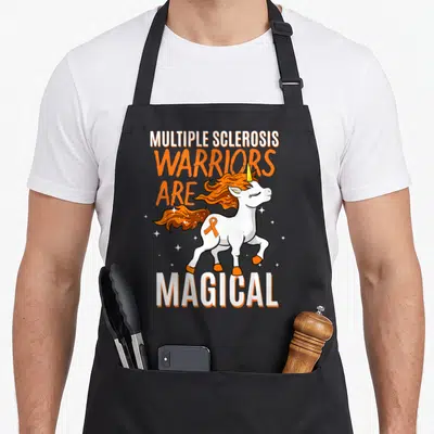 Niedliche unicorn apron - Cartoon white unicorn with orange mane and ribbon surrounded by stacked Multiple Sclerosis — shown on a black cotton canvas apron against a white background