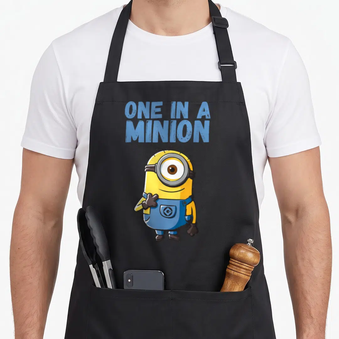 Despicable general apron - One In A Minion text above single-eyed yellow minion in blue overalls on grey background — shown on a black cotton canvas apron against a white background