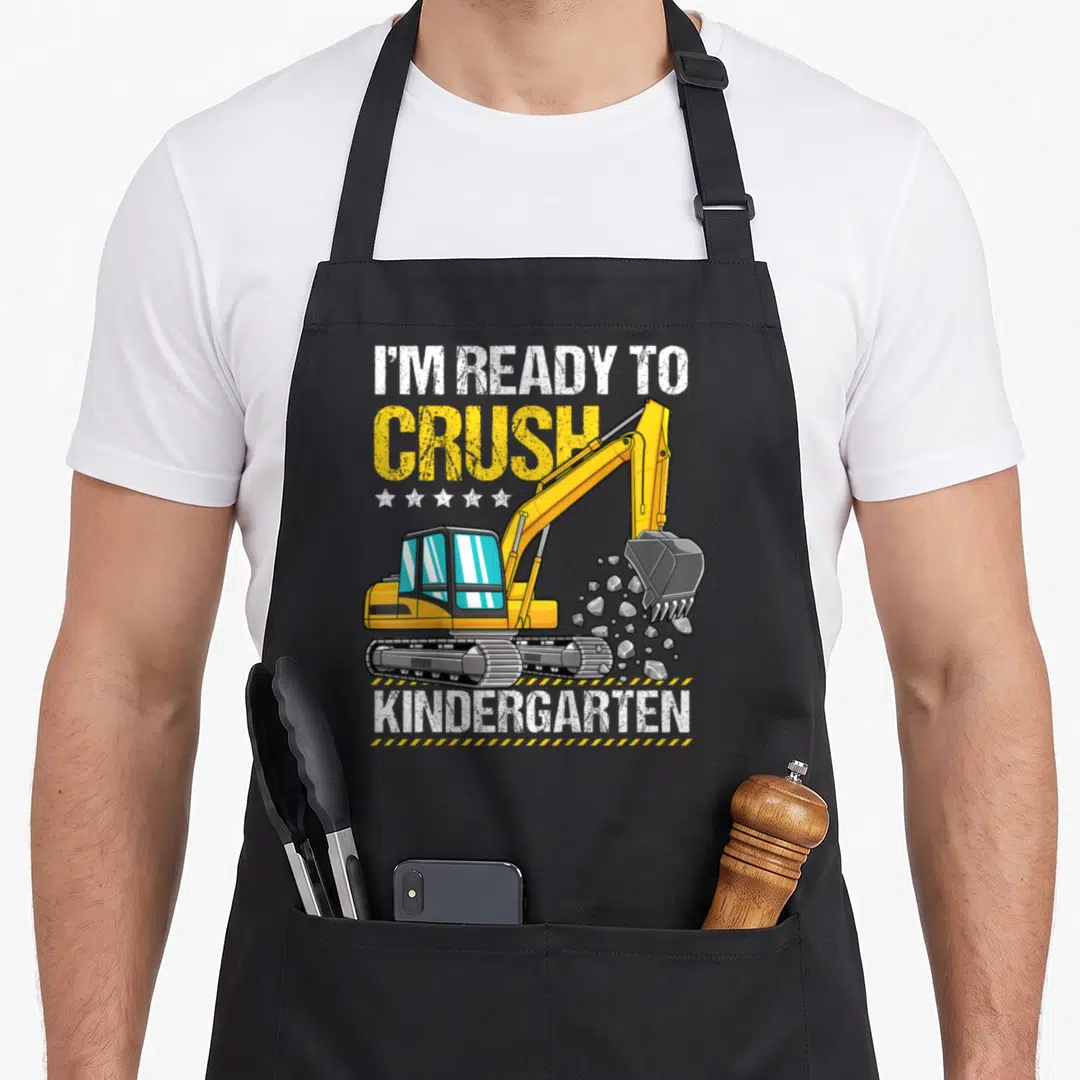 Ready kindergarten apron - Excavator digging rocks with I'm Ready To Crush Kindergarten text and stars — shown on a black cotton canvas apron against a white background