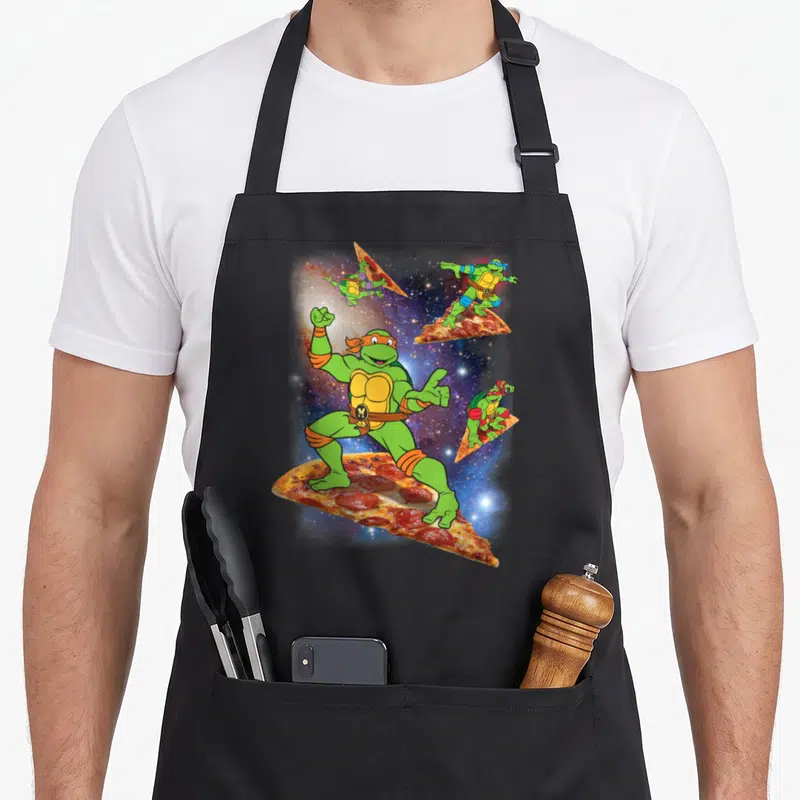 Cosmic pizza apron - Four green turtles surfing on pizza slices in space, wearing orange purple and blue gear — shown on a black cotton canvas apron against a white background