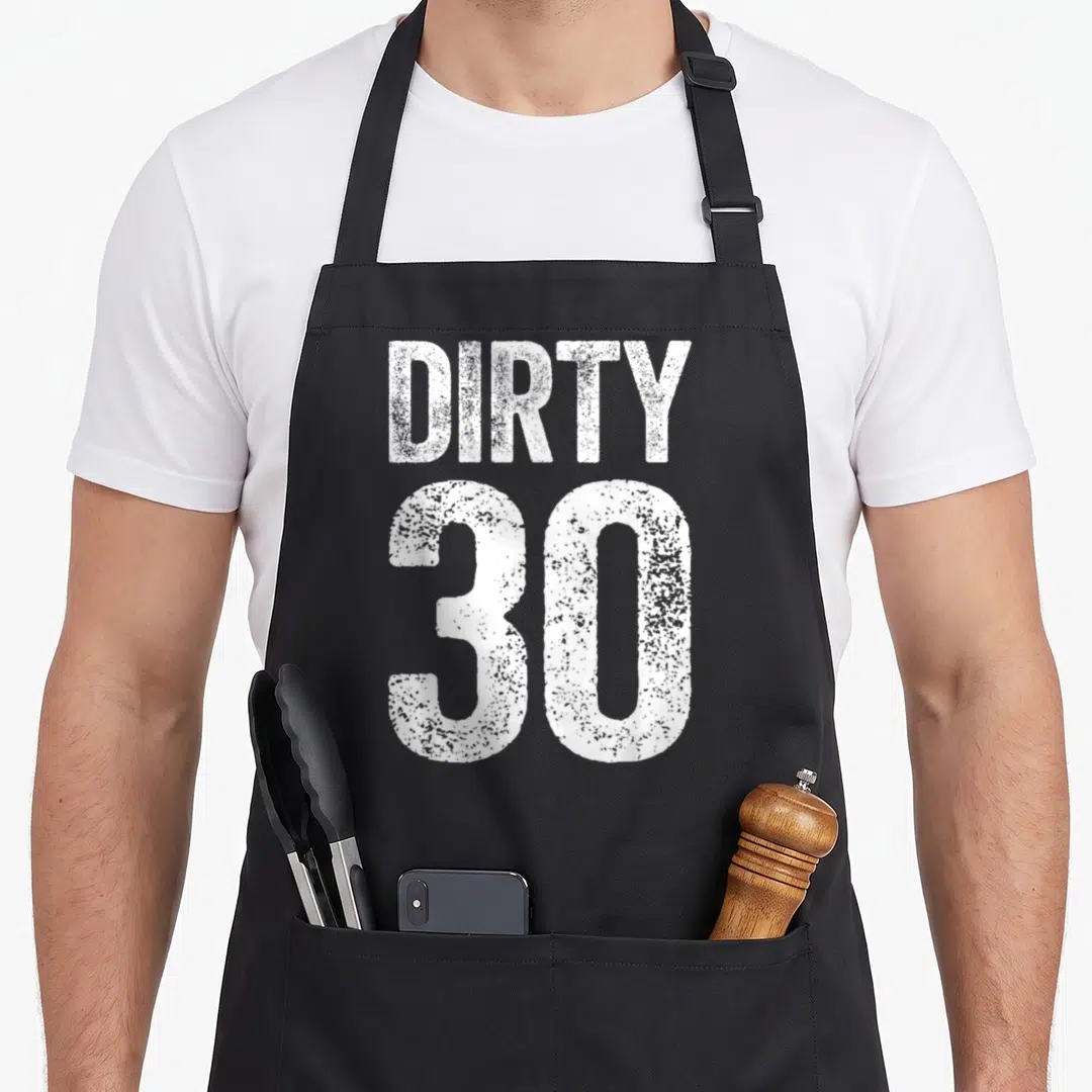 Thirty funny dirty aprons - White distressed typography reading DIRTY 30 on black fabric — shown on a black cotton canvas apron against a white background