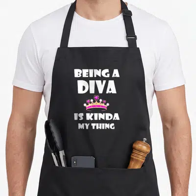 Funny diva birthday apron - White text reading Being a Diva Is Kinda My Thing with pink gold crown and hearts — shown on a black cotton canvas apron against a white background