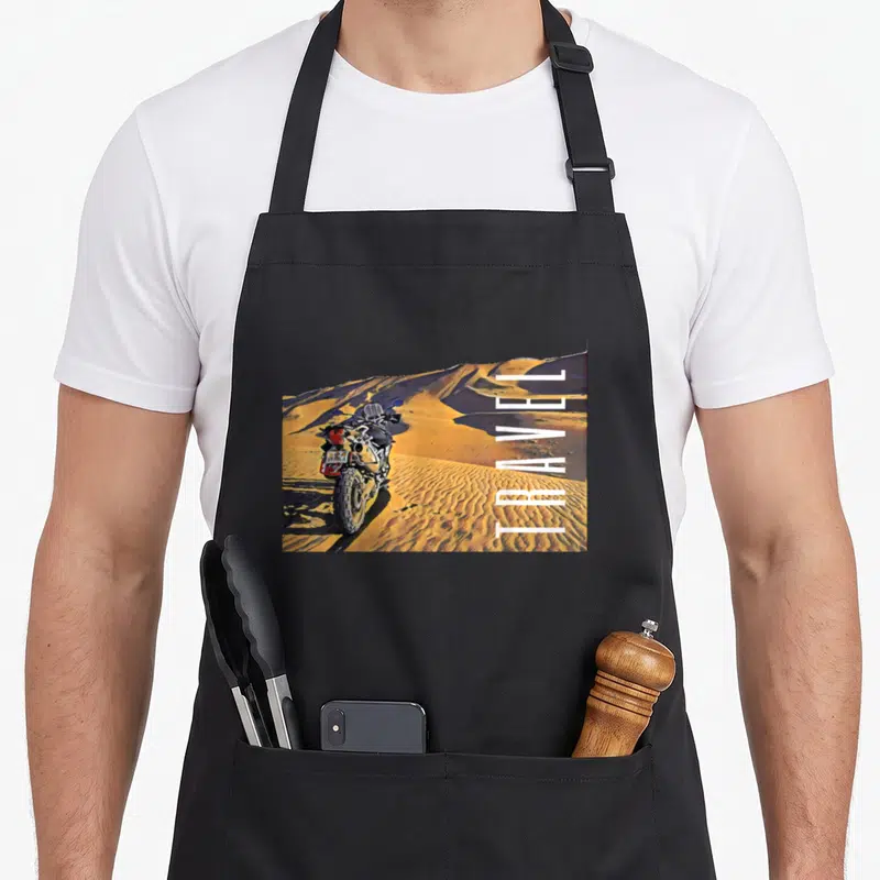 Motorcycle travel adventure bike apron - Vertical TRAVEL text beside a motorcycle on orange and yellow desert dunes — shown on a black cotton canvas apron against a white background