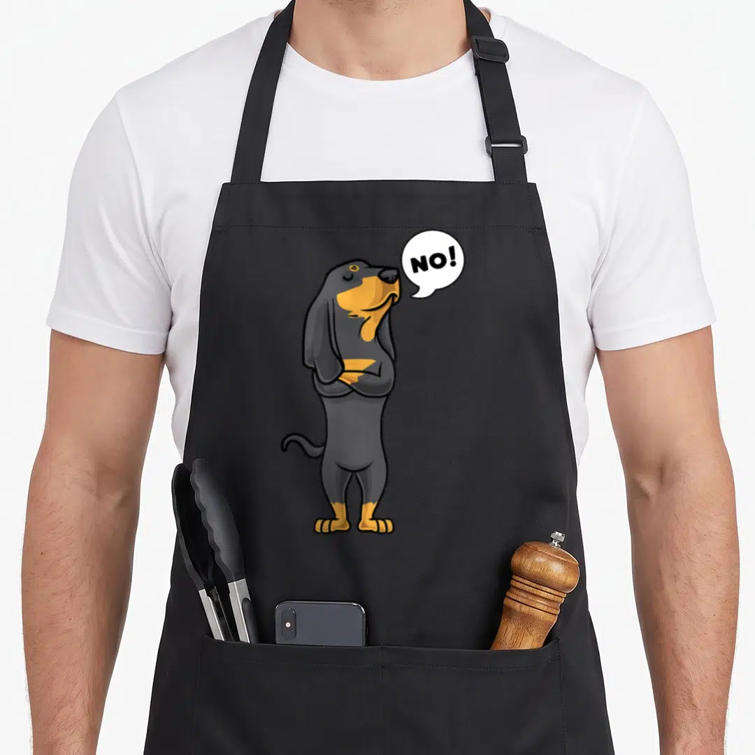 Stubborn coonhound dog apron - Cartoon black and tan dachshund standing with arms crossed next to a speech bubble — shown on a black cotton canvas apron against a white background