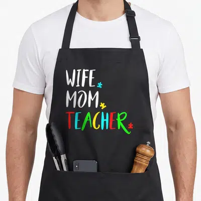 Autism best teacher apron - Stacked WIFE MOM TEACHER text with multicolored puzzle pieces — shown on a black cotton canvas apron against a white background