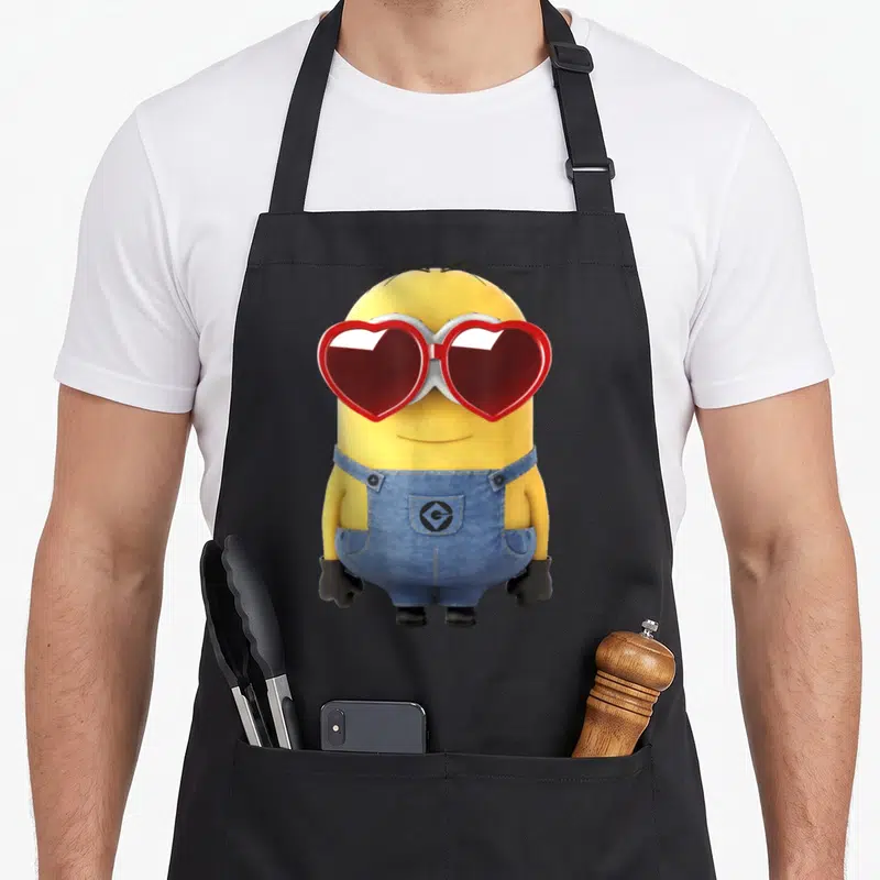Dave valentine's day apron - Yellow Minion wearing blue overalls and red heart-shaped sunglasses standing against black — shown on a black cotton canvas apron against a white background
