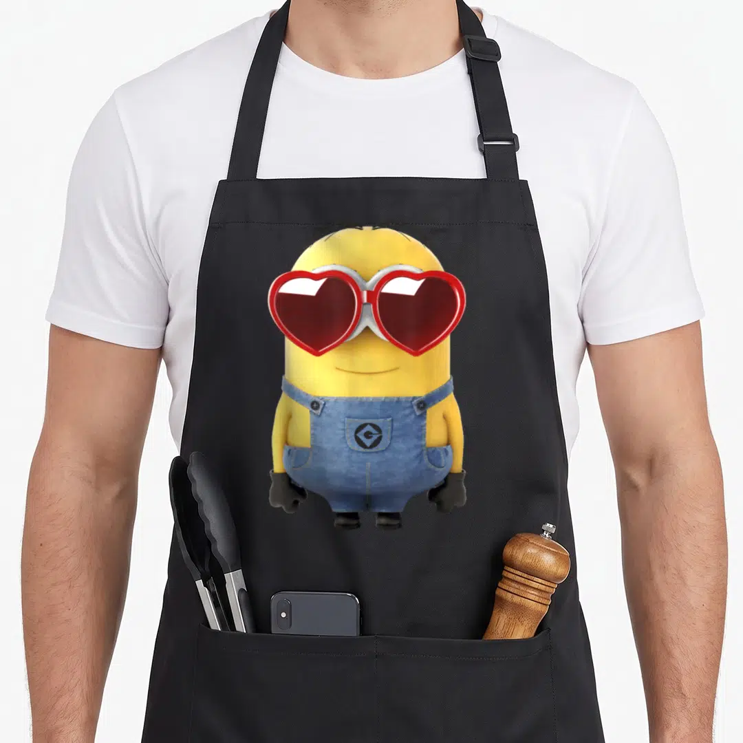 Dave valentine's day apron - Yellow Minion wearing blue overalls and red heart-shaped sunglasses standing against black — shown on a black cotton canvas apron against a white background