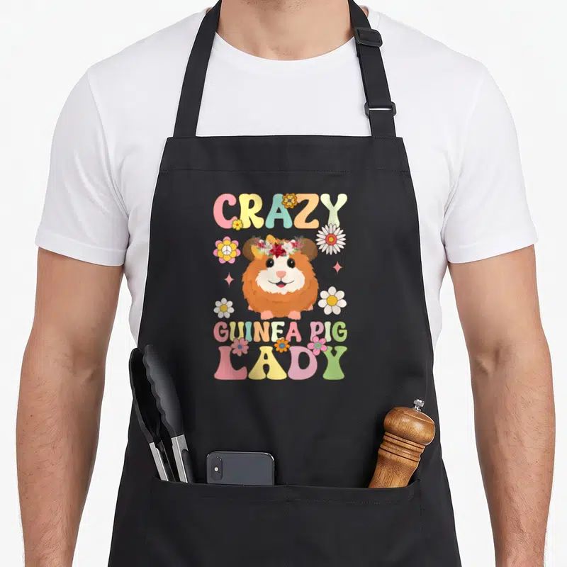 Cute groovy crazy guinea lady apron - Cartoon guinea pig with flower crown and pink yellow green orange bubble text — shown on a black cotton canvas apron against a white background