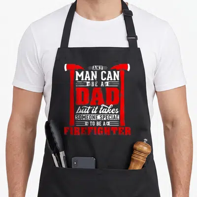 Special firefighter apron - Red and white stacked text reading any man can be a dad framed by two axes — shown on a black cotton canvas apron against a white background
