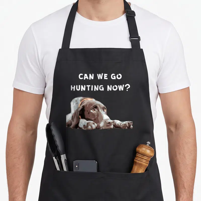 Hair pointer apron - Can We Go Hunting Now text above a brown and white dog lying down in cartoon style — shown on a black cotton canvas apron against a white background