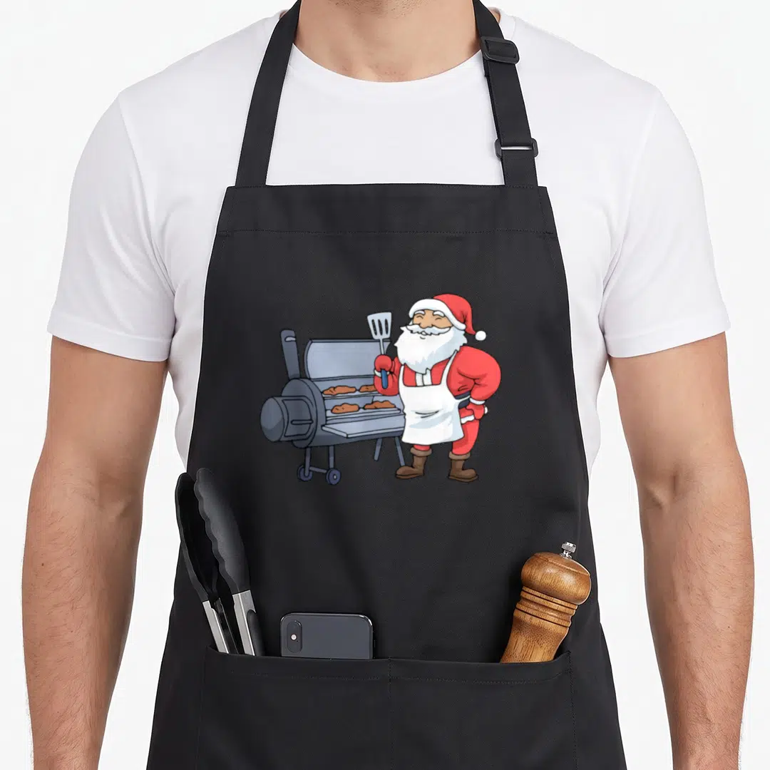 Barbecue santa claus apron - Cartoon Santa holding spatula next to grey smoker grill with brown meat cuts — shown on a black cotton canvas apron against a white background