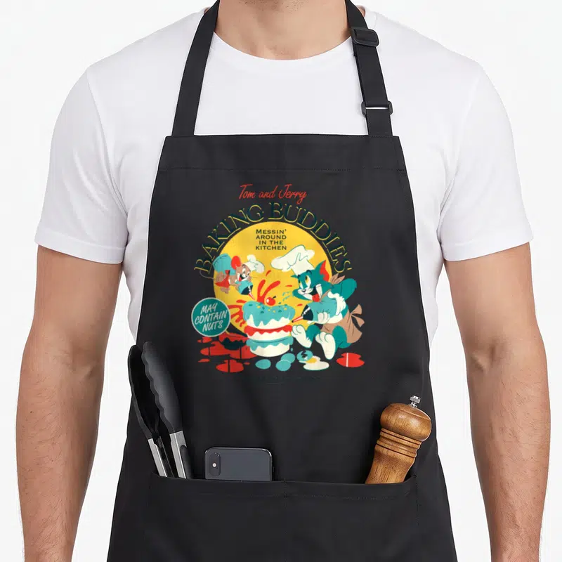 Cute buddy aprons - Tom and Jerry decorating a cake with red icing and arched Baking Buddies text — shown on a black cotton canvas apron against a white background