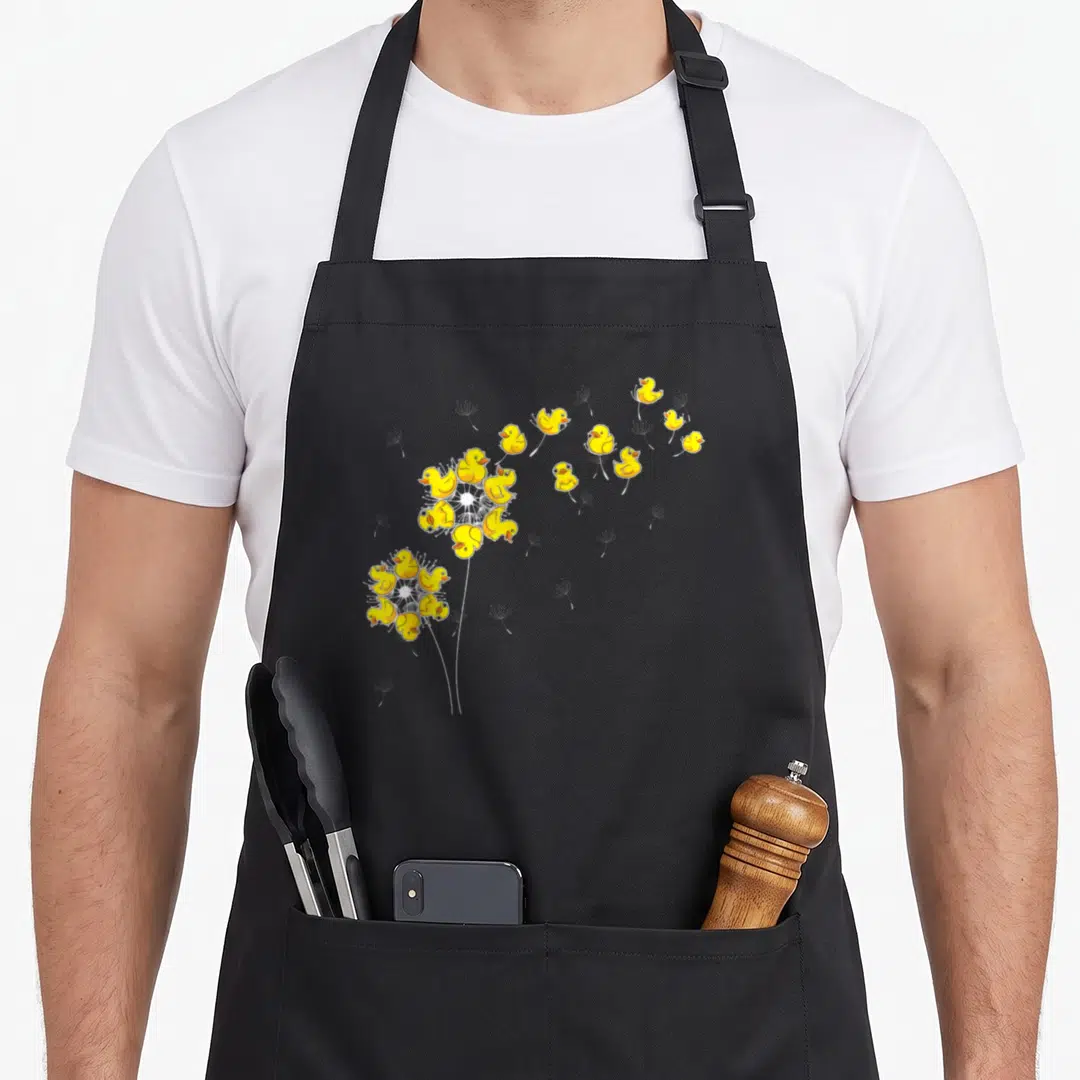 Dandelion duck apron - Yellow rubber ducks arranged as dandelion seeds blowing from white stems in cartoon style — shown on a black cotton canvas apron against a white background