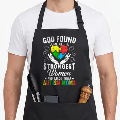 Autism mother's apron - Colorful puzzle heart cradled by hands with stacked distressed text reading God Found Some — shown on a black cotton canvas apron against a white background