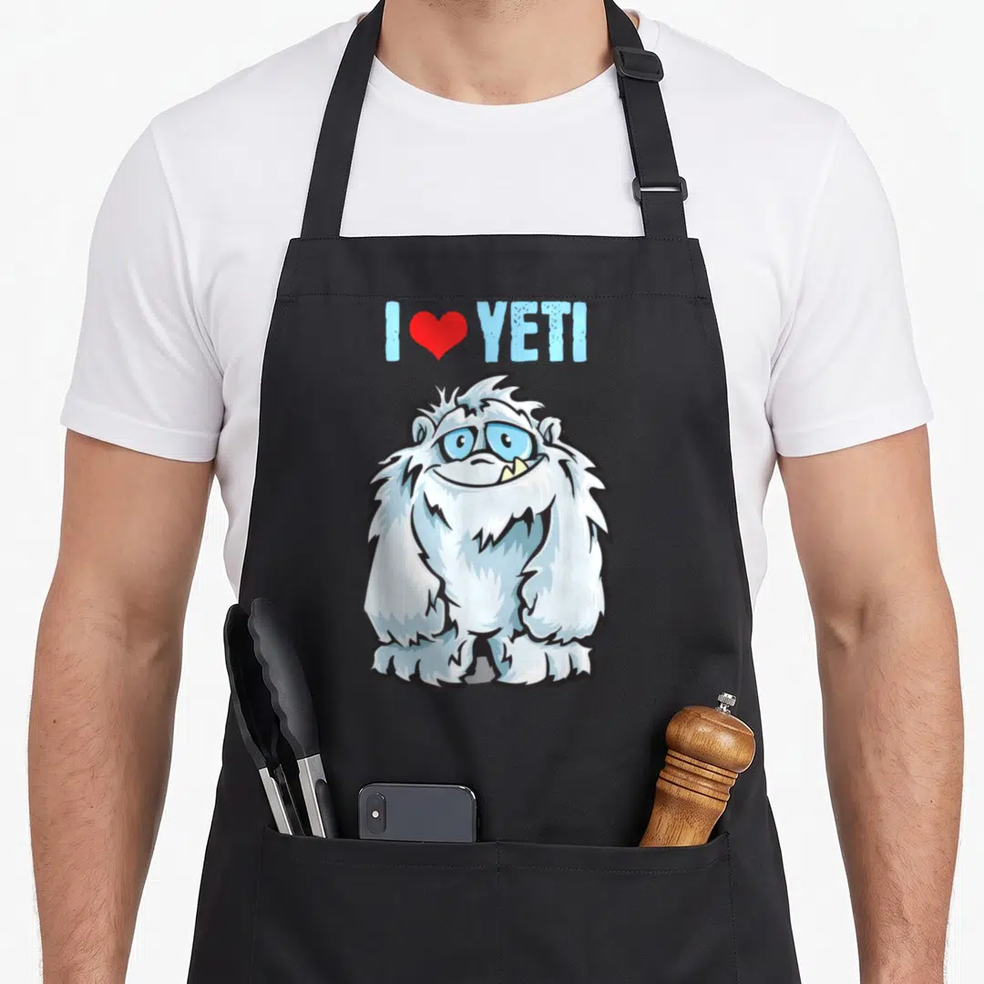 Cute yeti apron - I Heart YETI text with red heart above a fluffy white and light blue cartoon monster — shown on a black cotton canvas apron against a white background
