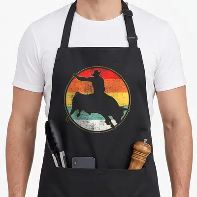 Bull cowboy apron - Grey cowboy silhouette riding bull over retro red orange yellow teal stripes — shown on a black cotton canvas apron against a white background