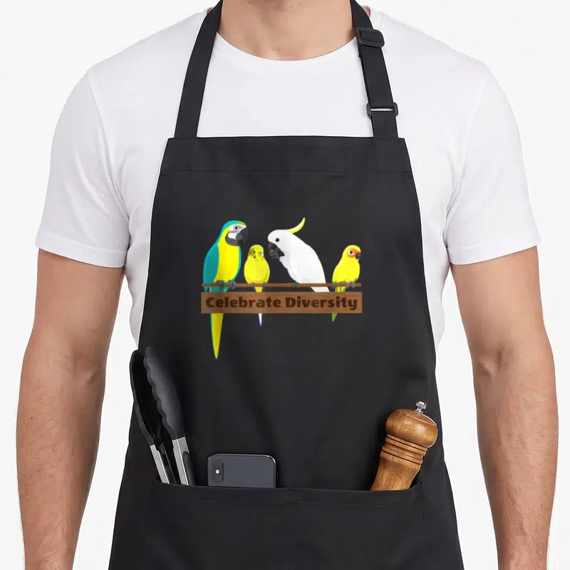 Celebrate parrot apron - Four parrots perched on a brown bar above a wooden sign reading Celebrate Diversity in cartoon — shown on a black cotton canvas apron against a white background