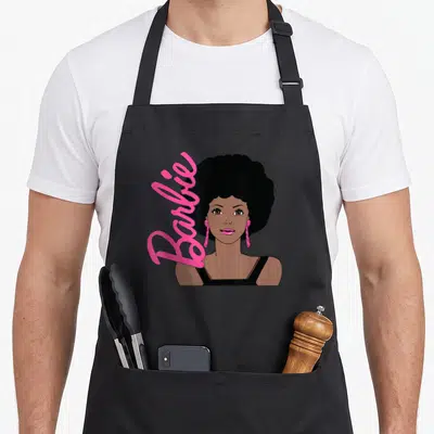Portrait afro apron - Cartoon woman with black afro and vertical pink Barbie text on brown skin — shown on a black cotton canvas apron against a white background