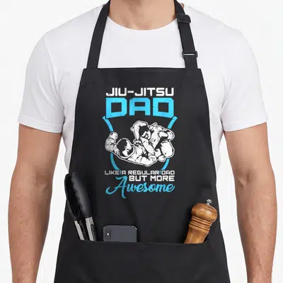 Jiu dance dad apron - Two grapplers in white gi inside blue shield with JIU-JITSU DAD text — shown on a black cotton canvas apron against a white background