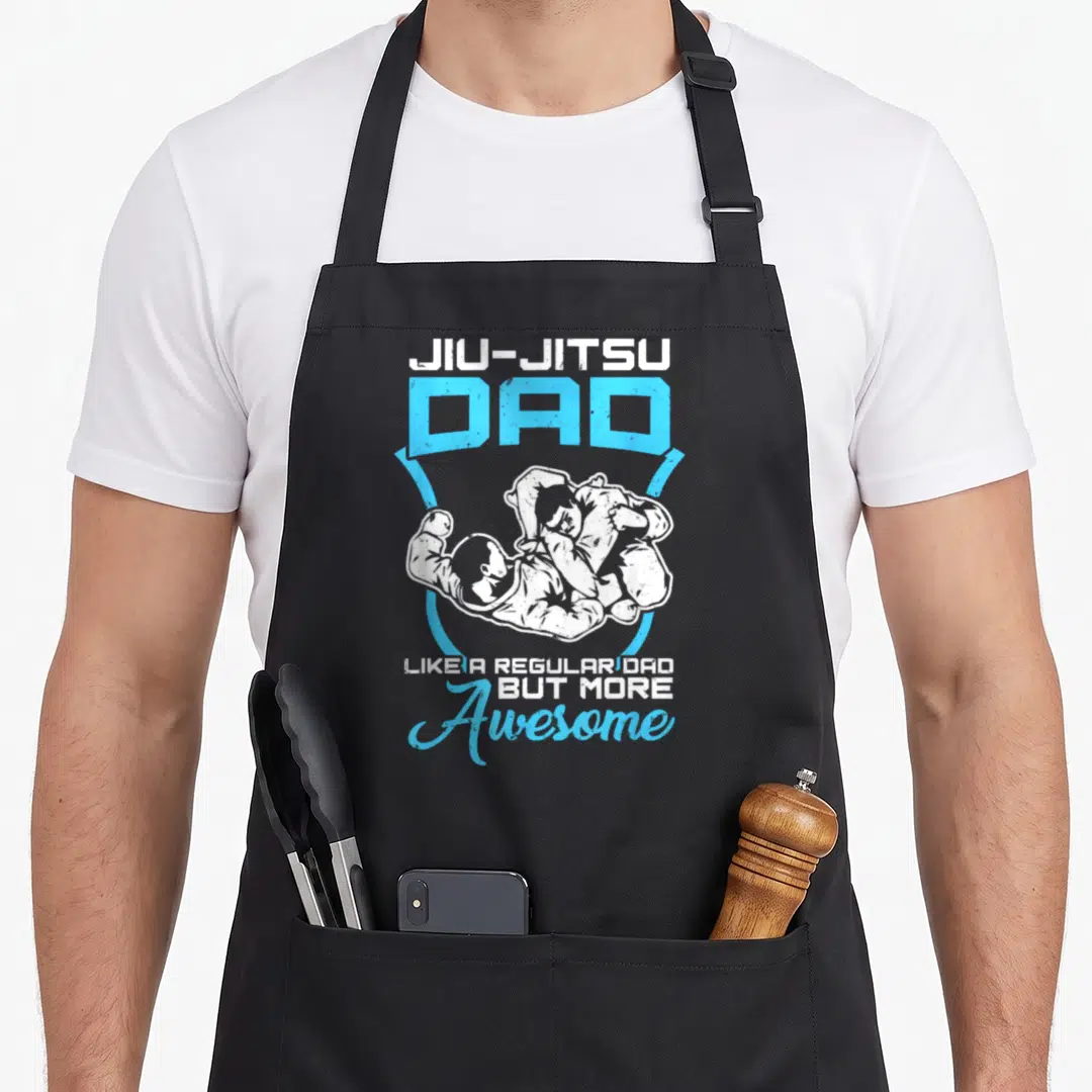 Jiu dance dad apron - Two grapplers in white gi inside blue shield with JIU-JITSU DAD text — shown on a black cotton canvas apron against a white background