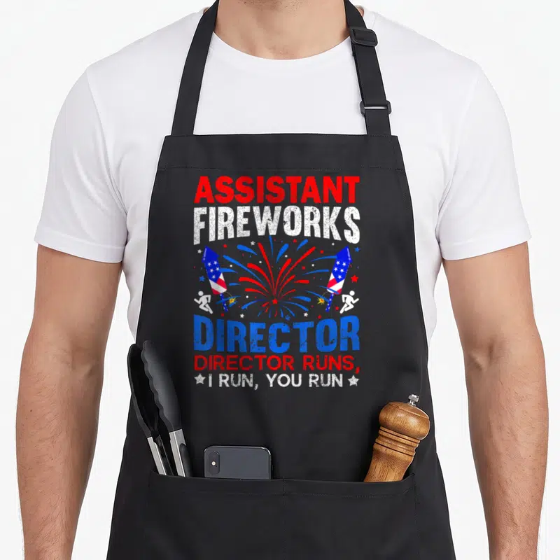 Assistant 4th of july apron - Distressed red white blue text with rockets and two running figures — shown on a black cotton canvas apron against a white background