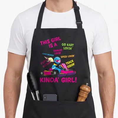 Funny kart racing apron - Cartoon driver in pink kart surrounded by colorful phrases about speed and rubber burnin' — shown on a black cotton canvas apron against a white background