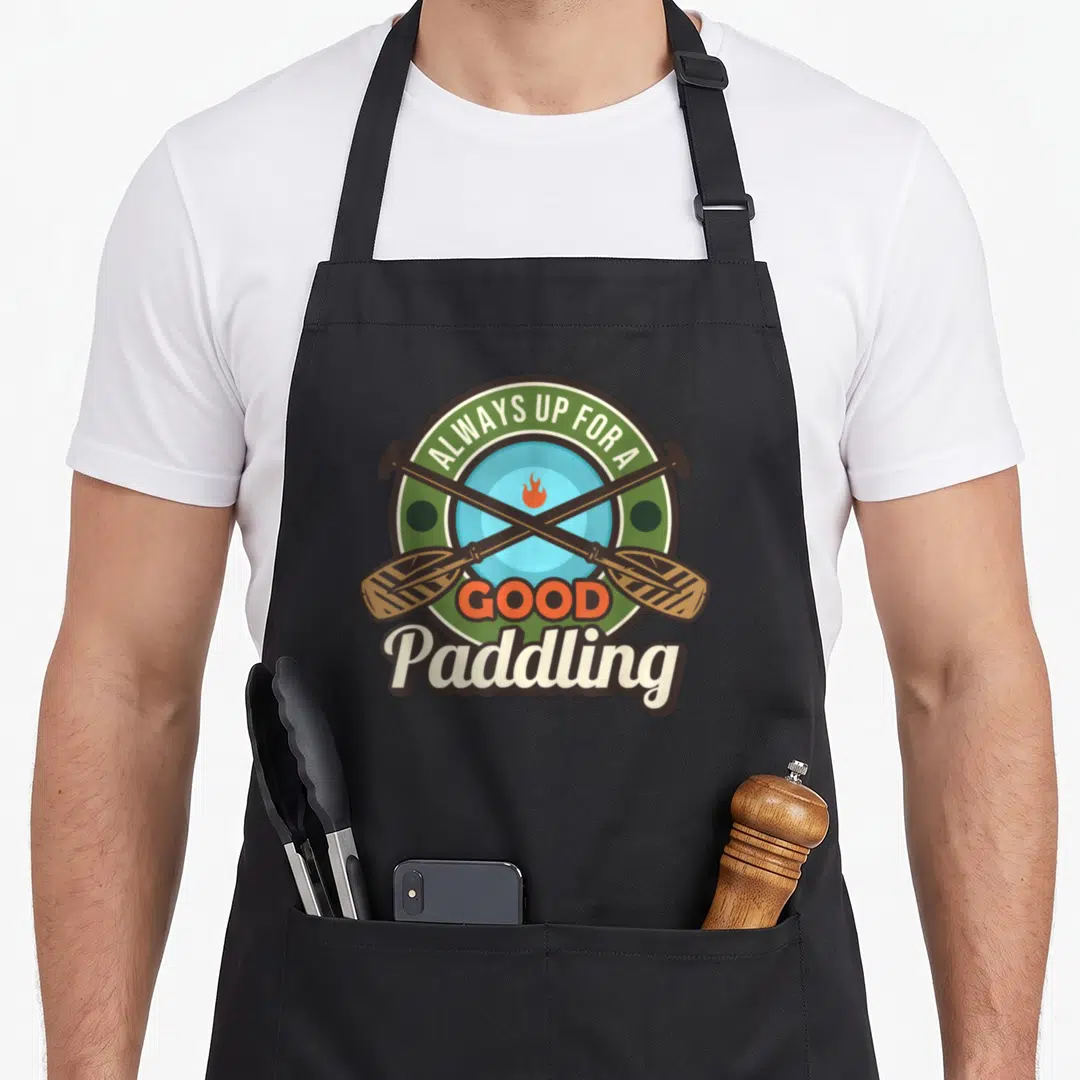 Alway paddleboard apron - Circular badge with crossed brown paddles, orange flame, and text ALWAYS UP FOR A GOOD — shown on a black cotton canvas apron against a white background
