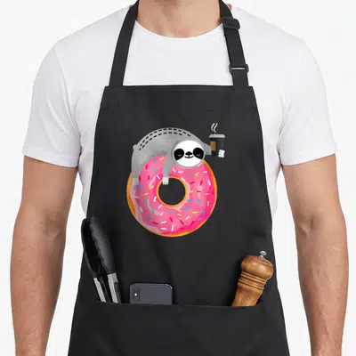 Donut sloth apron - Grey cartoon sloth on pink sprinkled donut holding black steaming coffee cup — shown on a black cotton canvas apron against a white background
