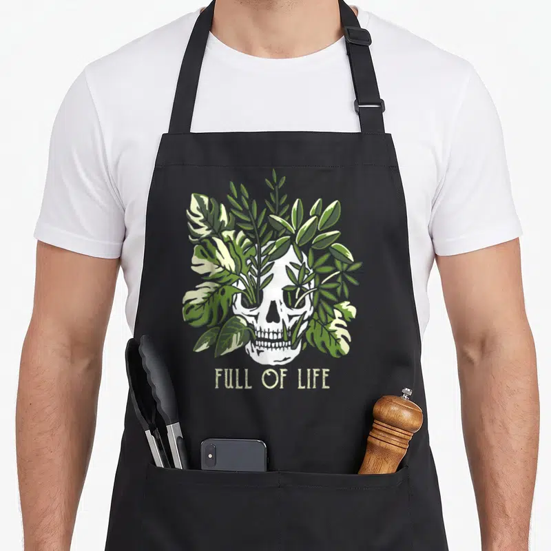 Gardening skull apron - White skull covered in green leaves with text FULL OF LIFE below — shown on a black cotton canvas apron against a white background