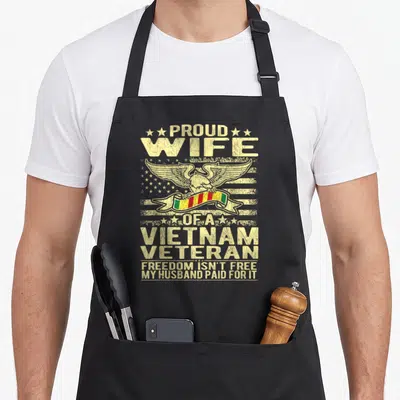 Free wife apron - Proud Wife Vietnam Veteran eagle holding red yellow green ribbon on distressed flag background — shown on a black cotton canvas apron against a white background